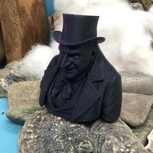 May include: A dark purple bust of a man in a top hat and formal wear. The sculpture is placed on rocks and oyster shells, with wood and cotton in the background. The bust has detailed facial features and clothing.