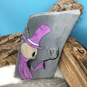 May include: Hand-painted gray rock art featuring a skeleton figure in a purple hat and coat, holding a snow globe with a red figure inside. The artwork includes a feather detail and is set against a blue background with wood accents.