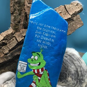 Pode incluir: Uma pedra azul pintada &agrave; m&atilde;o apresenta um personagem de desenho animado verde usando um cachecol listrado vermelho e branco. A pedra tem texto branco em alem&atilde;o, incluindo "Gl&uuml;hwein ist wie Tee nur lustiger". A pedra &eacute; exibida com madeira e outros elementos decorativos.