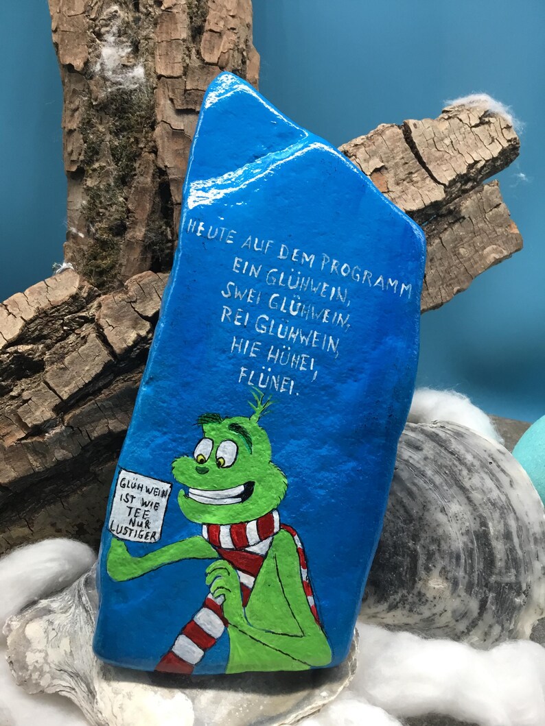 Pode incluir: Uma pedra azul pintada &agrave; m&atilde;o apresenta um personagem de desenho animado verde usando um cachecol listrado vermelho e branco. A pedra tem texto branco em alem&atilde;o, incluindo "Gl&uuml;hwein ist wie Tee nur lustiger". A pedra &eacute; exibida com madeira e outros elementos decorativos.