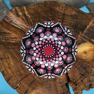 Op de afbeelding: Een ronde, zwarte steen beschilderd met een roze en wit mandala-ontwerp. De steen ligt op een houten oppervlak.
