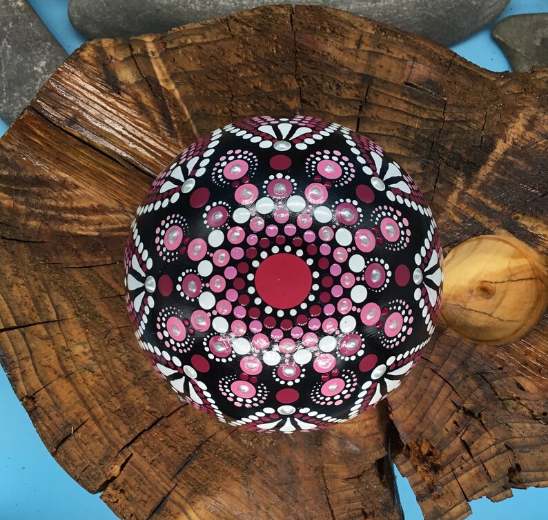 Op de afbeelding: Een ronde, zwarte steen beschilderd met een roze en wit mandala-ontwerp. De steen ligt op een houten oppervlak.
