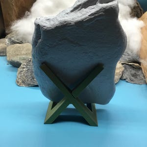 May include: A large, irregular, light gray stone with a textured, matte surface is held upright by a dark olive green, X-shaped plastic stand. The stone features natural crevices and a jagged top edge, resting securely in the center of the stand.