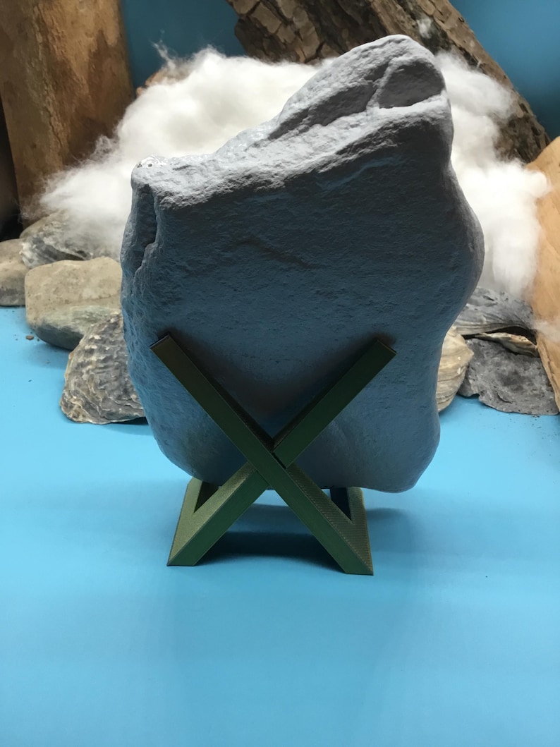 May include: A large, irregular, light gray stone with a textured, matte surface is held upright by a dark olive green, X-shaped plastic stand. The stone features natural crevices and a jagged top edge, resting securely in the center of the stand.