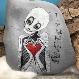 May include: Hand-painted gray rock art featuring a skeletal figure holding a red heart. The figure has a white skull-like head with large black eyes. Text on the rock reads "I do find most people to be awfully boring."