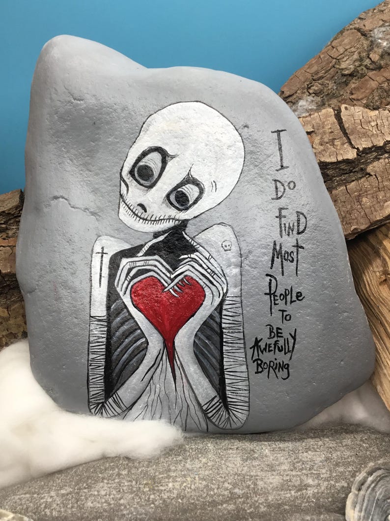 May include: Hand-painted gray rock art featuring a skeletal figure holding a red heart. The figure has a white skull-like head with large black eyes. Text on the rock reads "I do find most people to be awfully boring."