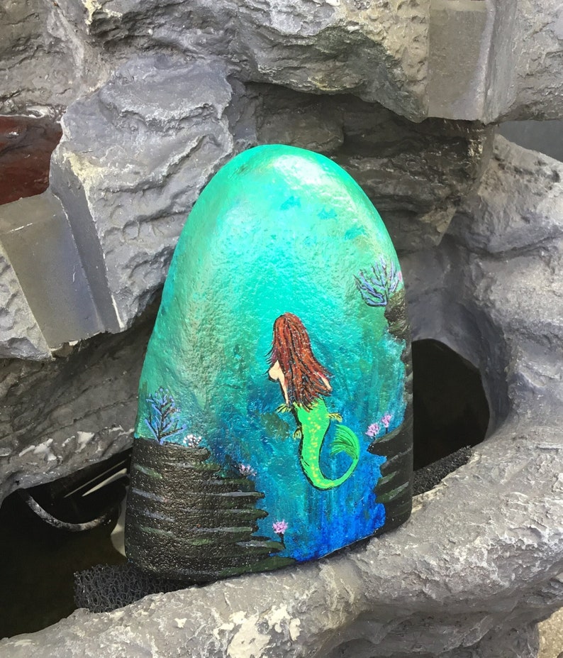 May include: Hand-painted rock featuring a mermaid design. The mermaid is green and has long red hair. The background is blue and green with black rocks and pink flowers.