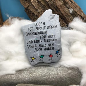May include: A light gray painted stone featuring black hand-lettered German text that reads: LEBEN IST NICHT GENUG SONNENSCHEIN FREIHEIT UND EINEN KLEINEN VOGEL MUSS MAN AUCH HABEN. Below the text are four small, colorful bird illustrations in yellow, green, red, and blue.