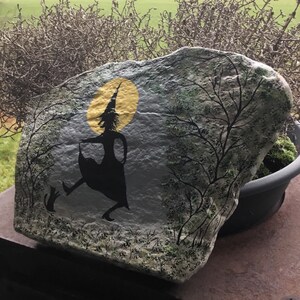 May include: A painted rock with a silhouette of a witch flying with a cat. The witch is wearing a pointed hat and a long dress. The background is a yellow moon and a dark forest.