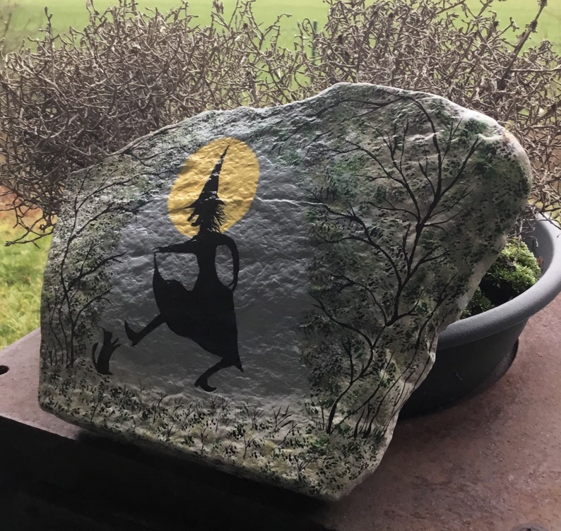 May include: A painted rock with a silhouette of a witch flying with a cat. The witch is wearing a pointed hat and a long dress. The background is a yellow moon and a dark forest.