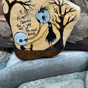 Peut inclure: Galet peint &agrave; la main avec une sc&egrave;ne fantaisiste. Il repr&eacute;sente une silhouette noire, deux orbes flottants et des arbres d&eacute;nud&eacute;s. Le texte "I Always Searched for a Way Out of The Cruel World" est &eacute;crit sur le galet. La palette de couleurs comprend beige, marron, noir et blanc.