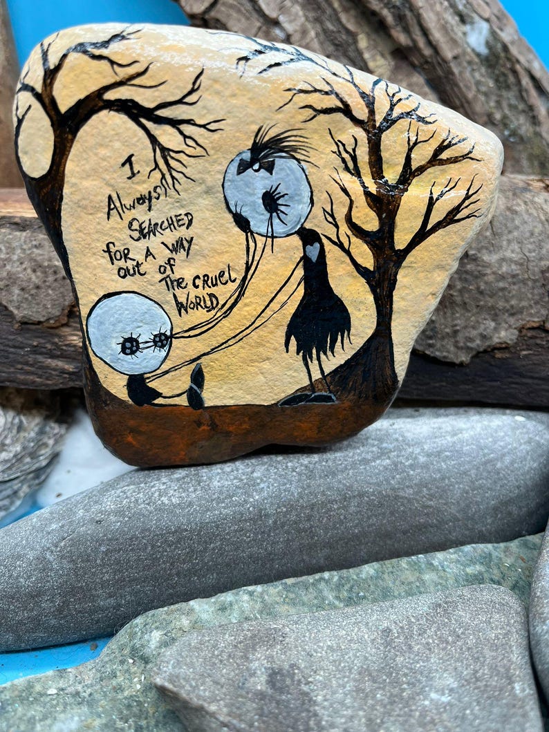 Peut inclure: Galet peint &agrave; la main avec une sc&egrave;ne fantaisiste. Il repr&eacute;sente une silhouette noire, deux orbes flottants et des arbres d&eacute;nud&eacute;s. Le texte "I Always Searched for a Way Out of The Cruel World" est &eacute;crit sur le galet. La palette de couleurs comprend beige, marron, noir et blanc.