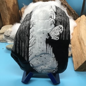 May include: A hand-painted rock depicting a black and white forest scene with a fairy. The fairy has a detailed wing design. The rock is displayed on a blue stand. The artwork depicts a forest with trees and stars.