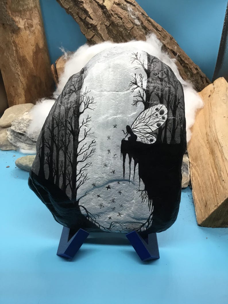 May include: A hand-painted rock depicting a black and white forest scene with a fairy. The fairy has a detailed wing design. The rock is displayed on a blue stand. The artwork depicts a forest with trees and stars.