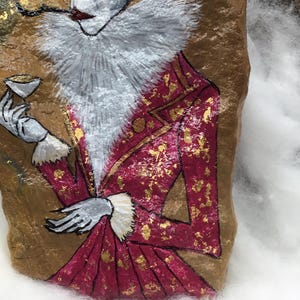 Op de afbeelding: Een beschilderde steen met een figuur in een rood en goudkleurig jasje, een witte gefranjerde kraag en een klein kopje. De figuur heeft een bleek gezicht, donker haar en rookt een pijp. De achtergrond is goudkleurig.