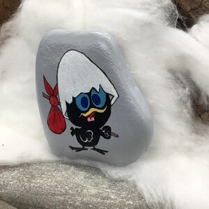 K&ouml;nnte beinhalten: Ein handbemalter, grauer Stein mit einem Cartoon-K&uuml;ken. Das K&uuml;ken ist schwarz mit blauen Augen, einem gelben Schnurrbart und einer roten Tasche. Das K&uuml;ken schl&uuml;pft aus einer wei&szlig;en Eierschale.