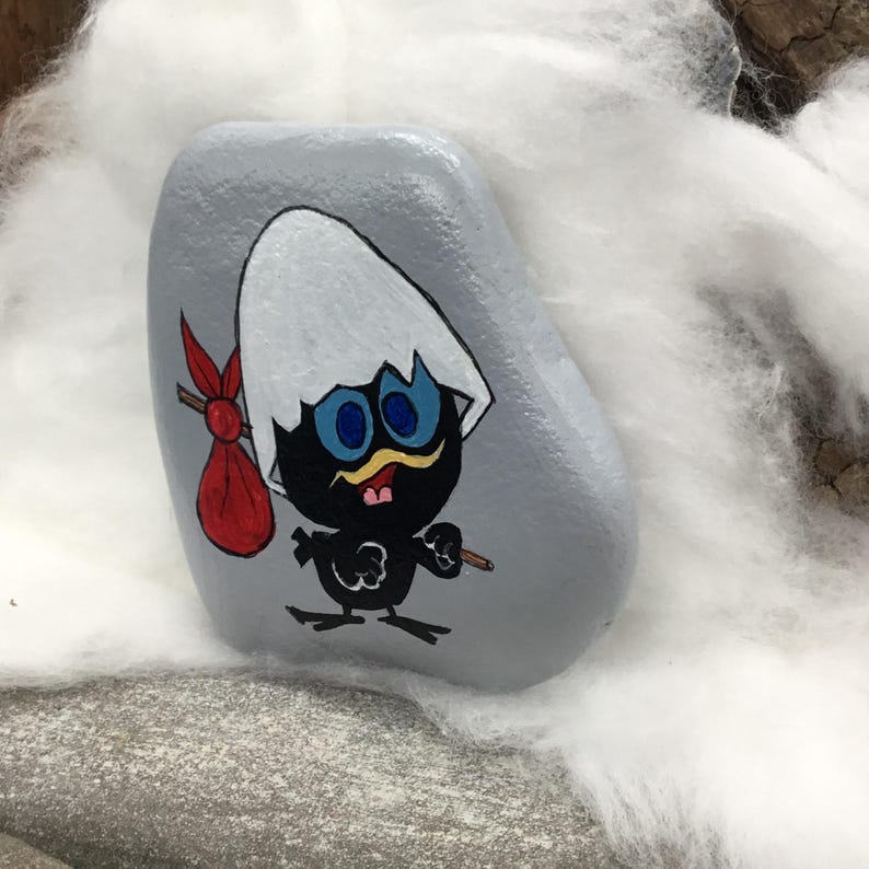 K&ouml;nnte beinhalten: Ein handbemalter, grauer Stein mit einem Cartoon-K&uuml;ken. Das K&uuml;ken ist schwarz mit blauen Augen, einem gelben Schnurrbart und einer roten Tasche. Das K&uuml;ken schl&uuml;pft aus einer wei&szlig;en Eierschale.