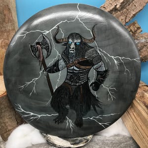 May include: A circular, grey painted stone depicts a mythical beast with bull horns, blue eyes, and a long black beard. The creature holds an axe and is surrounded by white lightning bolts. The artwork is detailed and has a fantasy theme.