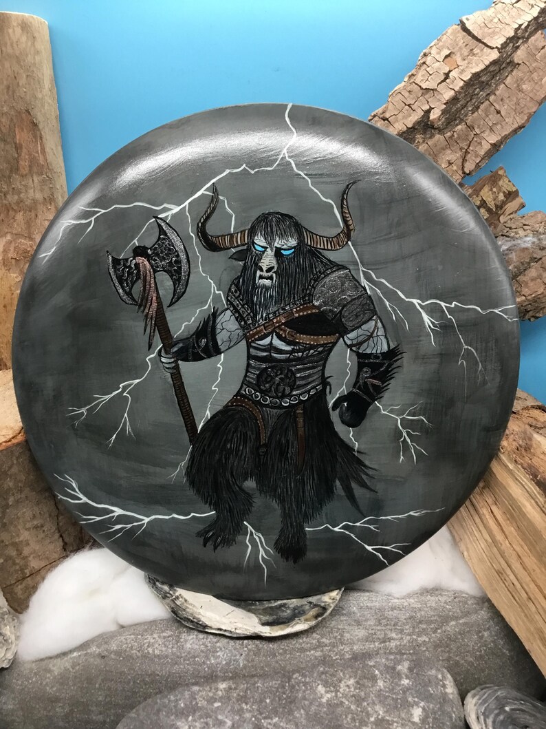 May include: A circular, grey painted stone depicts a mythical beast with bull horns, blue eyes, and a long black beard. The creature holds an axe and is surrounded by white lightning bolts. The artwork is detailed and has a fantasy theme.
