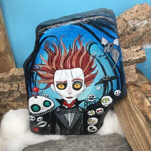 Pu&ograve; includere: Roccia dipinta a mano raffigurante un personaggio con capelli rossi e occhi gialli, vestito con un abito nero con papillon rosso. Lo sfondo blu presenta pipistrelli e altre creature. Un'opera d'arte unica.