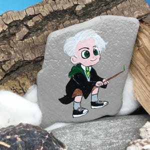 May include: Hand-painted rock art featuring a cartoon character with white hair, green eyes, and a green and black robe. The character is holding a wand. The rock is gray and sits on a natural background.