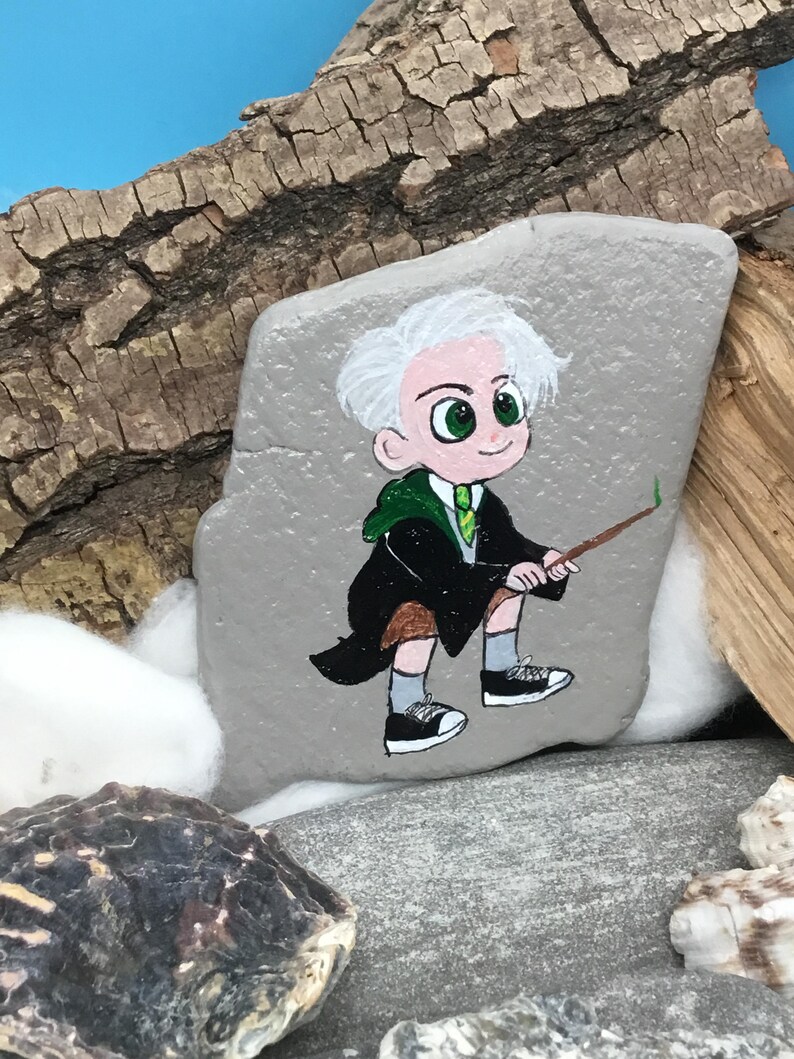 May include: Hand-painted rock art featuring a cartoon character with white hair, green eyes, and a green and black robe. The character is holding a wand. The rock is gray and sits on a natural background.