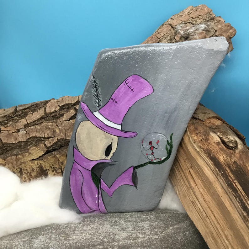 May include: A hand-painted, gray stone features a whimsical illustration of a skeleton figure in a purple top hat and coat, holding a crystal ball. The artwork includes a feather and a small figure inside the ball. The stone is set against a blue backdrop.