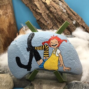 May include: A light blue painted rock featuring a hand-painted illustration of a character with red hair in pigtails, wearing a yellow dress, blue polka-dot shorts, and black boots with yellow and black striped stockings, displayed on a green stand.