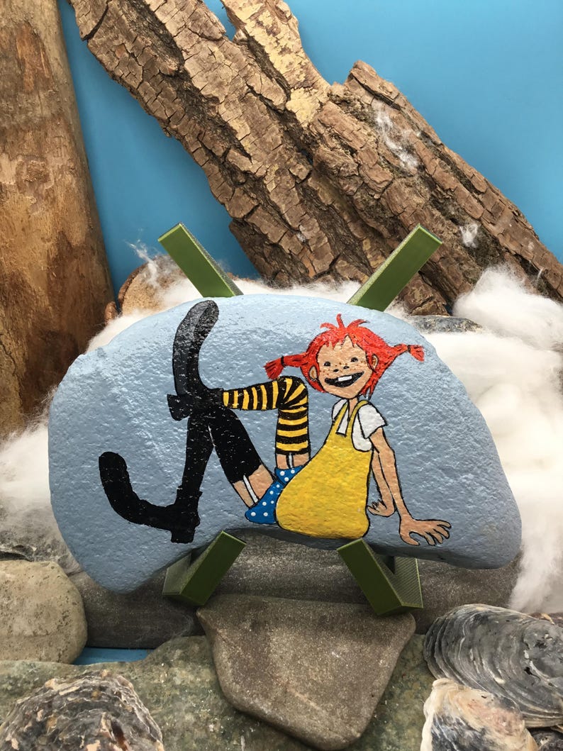 May include: A light blue painted rock featuring a hand-painted illustration of a character with red hair in pigtails, wearing a yellow dress, blue polka-dot shorts, and black boots with yellow and black striped stockings, displayed on a green stand.