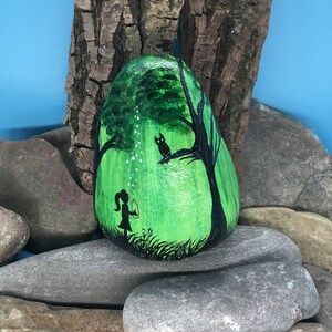 K&ouml;nnte beinhalten: Handbemalter Stein mit der Darstellung eines M&auml;dchens, das Seifenblasen in einem gr&uuml;nen Wald bl&auml;st, mit einer schwarzen Katze, die auf einem Baumzweig sitzt.