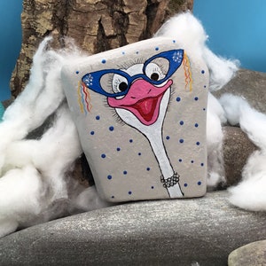 May include: Hand-painted rock art of an ostrich wearing blue glasses and a pearl necklace. The rock is painted with a light grey background and blue polka dots.