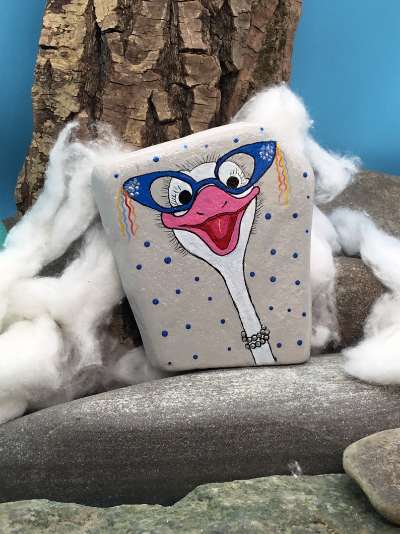 May include: Hand-painted rock art of an ostrich wearing blue glasses and a pearl necklace. The rock is painted with a light grey background and blue polka dots.
