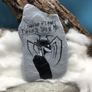 May include: A light gray painted rock featuring a black ink illustration of a stylized figure with long, spindly limbs and a white eye. The text "I DON'T LIVE IN DARKNESS DARKNESS LIVES IN ME" is written in black ink above the figure on the rock surface.