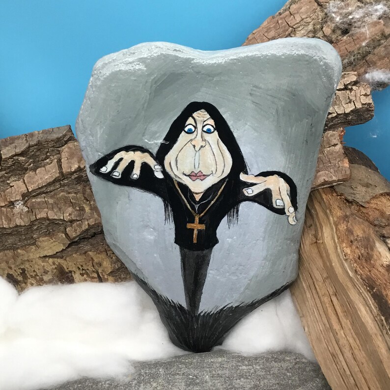 May include: Hand-painted rock art depicting a caricature of a person in a black robe and cross necklace. The gray rock has a painted face and arms. The background includes wood and a blue sky.