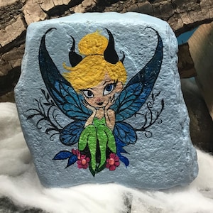 May include: Hand-painted rock art featuring a fairy with blue wings, green dress, and yellow hair. The fairy has black horns and is sitting on a flower.