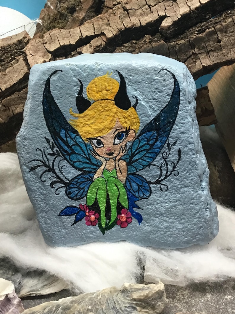 May include: Hand-painted rock art featuring a fairy with blue wings, green dress, and yellow hair. The fairy has black horns and is sitting on a flower.