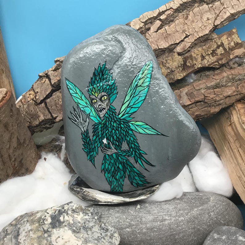 May include: A hand-painted gray rock features a detailed illustration of a fairy with green and teal wings and clothing. The fairy has a stern expression and is holding a small white object. The rock is surrounded by natural elements.