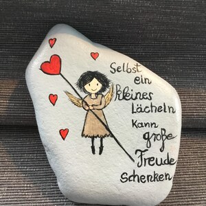 K&ouml;nnte beinhalten: Ein bemalter Stein mit hellblauem Hintergrund zeigt einen Cartoon-Engel, der einen herzf&ouml;rmigen Zauberstab h&auml;lt. Rote Herzen sind um den Engel verstreut. Der deutsche Text lautet: &bdquo;Selbst ein kleines L&auml;cheln kann gro&szlig;e Freude schenken&ldquo;.