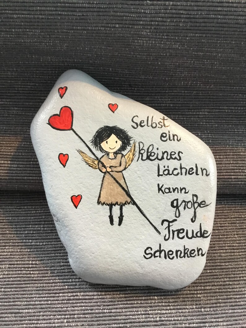 K&ouml;nnte beinhalten: Ein bemalter Stein mit hellblauem Hintergrund zeigt einen Cartoon-Engel, der einen herzf&ouml;rmigen Zauberstab h&auml;lt. Rote Herzen sind um den Engel verstreut. Der deutsche Text lautet: &bdquo;Selbst ein kleines L&auml;cheln kann gro&szlig;e Freude schenken&ldquo;.
