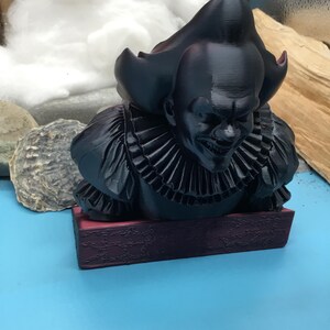 May include: A black bust of a menacing clown with a ruffled collar and a sinister grin. The figure is mounted on a burgundy base. The clown's hair is styled in a tall, pointed shape.