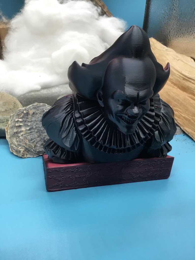 May include: A black bust of a menacing clown with a ruffled collar and a sinister grin. The figure is mounted on a burgundy base. The clown's hair is styled in a tall, pointed shape.