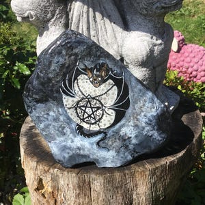 May include: A hand-painted, dark gray rock featuring a black bat with white accents and a pentagram design. The bat has a detailed face and is surrounded by a decorative chain. The rock is displayed on a wooden stump.