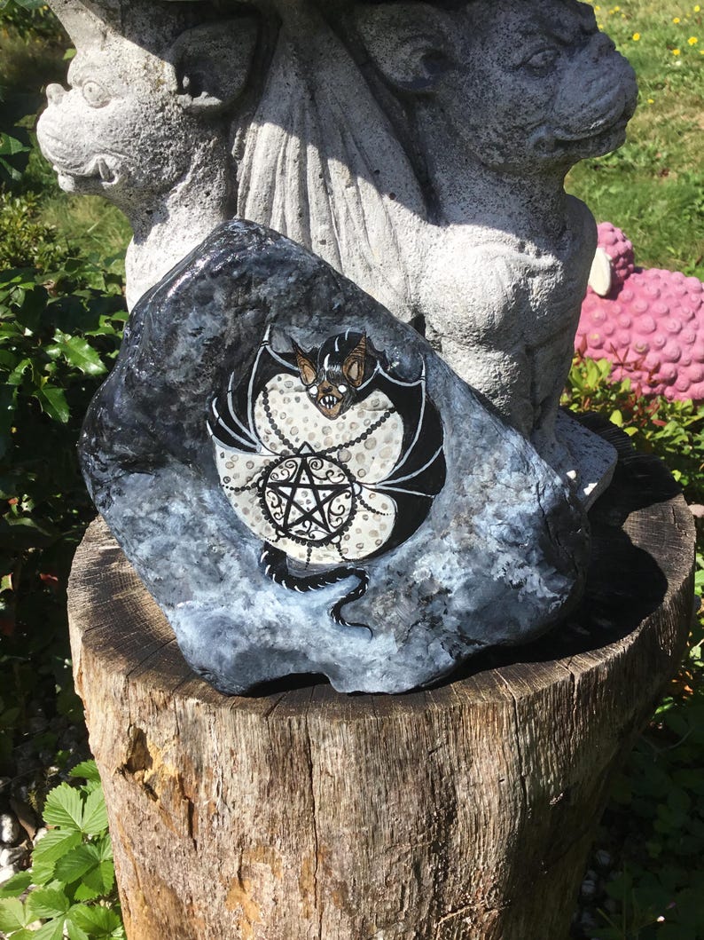 May include: A hand-painted, dark gray rock featuring a black bat with white accents and a pentagram design. The bat has a detailed face and is surrounded by a decorative chain. The rock is displayed on a wooden stump.
