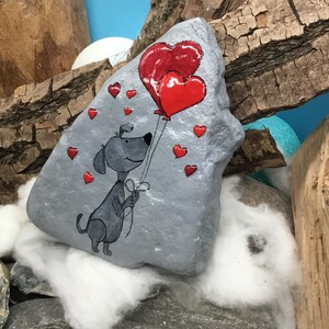 May include: A hand-painted gray rock with a cartoon dog holding red heart-shaped balloons. The rock is surrounded by small red hearts.
