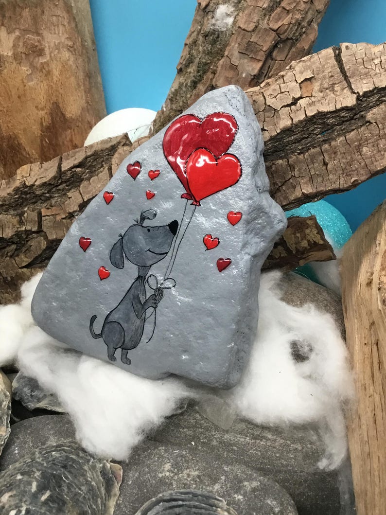 May include: A hand-painted gray rock with a cartoon dog holding red heart-shaped balloons. The rock is surrounded by small red hearts.