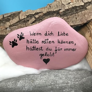 Può includere: Una pietra dipinta di rosa con impronte di zampe nere e il testo "Wenn dich Liebe hätte retten können, hättest du für immer gelebt" e un cuore nero. La pietra è appoggiata su un letto di lanugine bianca.