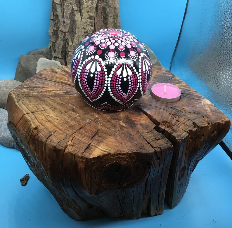 Op de afbeelding: Een ronde houten bol met een geschilderd roze en zwart mandala ligt op een rustieke houten stronk met een roze kaars in een glazen houder.