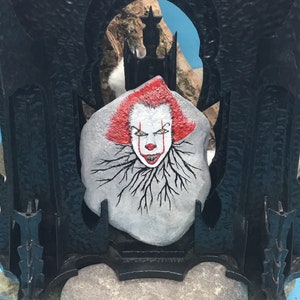 May include: A painted rock with a scary clown face, red hair, and black roots. The rock is set in a black metal frame with a gothic design.