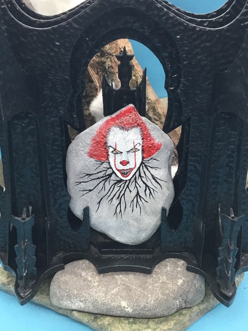 May include: A painted rock with a scary clown face, red hair, and black roots. The rock is set in a black metal frame with a gothic design.