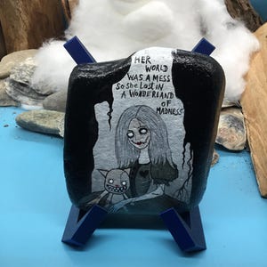 May include: A painted rock featuring a grayscale illustration of a girl and a creature, set against a dark backdrop. The text on the rock reads "Her world was a mess so she lost in a wonderland of madness." The rock is displayed on a blue stand.